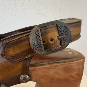 May include: Close-up of a brown leather sandal with a wooden platform heel. The sandal features a buckle with the word "Skocca" and decorative metal studs. The leather has a distressed appearance.