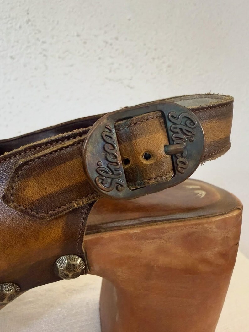 May include: Close-up of a brown leather sandal with a wooden platform heel. The sandal features a buckle with the word "Skocca" and decorative metal studs. The leather has a distressed appearance.