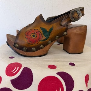 May include: Brown leather clogs with a floral design. The open-toe sandals feature a wooden platform and heel, a buckle closure, and a painted red rose with green leaves. The clogs are adorned with metal studs around the sole.