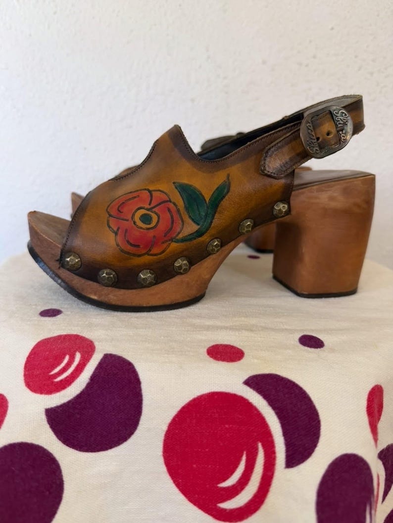 May include: Brown leather clogs with a floral design. The open-toe sandals feature a wooden platform and heel, a buckle closure, and a painted red rose with green leaves. The clogs are adorned with metal studs around the sole.