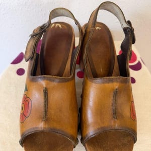 May include: A pair of vintage brown leather clogs with open toes and ankle straps. Each clog features a red and green floral design on the side. The wooden soles and leather straps are secured with buckles.