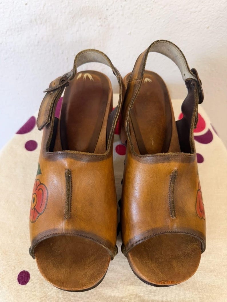 May include: A pair of vintage brown leather clogs with open toes and ankle straps. Each clog features a red and green floral design on the side. The wooden soles and leather straps are secured with buckles.