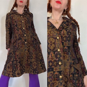 Puede incluir: Un abrigo vintage con un diseño estampado en tonos marrón, negro y verde oliva. El abrigo presenta botones dorados, bolsillos y un cuello ancho. El abrigo llega a la rodilla y tiene mangas largas.