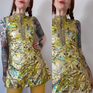May include: A vintage tunic top with a high collar and gold trim. The top features a floral pattern in gold, green, and blue. The tunic has a slight A-line shape and is made of a shiny, textured fabric. The model is wearing yellow pants.