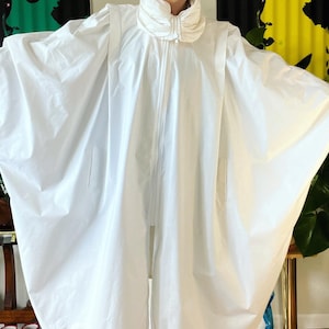 Puede incluir: Una chaqueta blanca, de gran tamaño,  y abultada con un cuello alto y cierre de cremallera. La chaqueta tiene una silueta suelta y fluida y está hecha de una tela ligera.