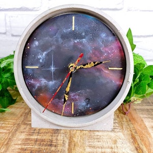 May include: A gray concrete clock with a galaxy-themed face. The clock face features a dark blue background with white and pink clouds, gold stars, and gold-colored hands. The clock is sitting on a wooden surface.