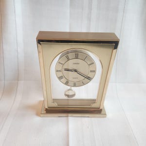 May include: A gold-toned quartz clock with a rectangular frame and a clear glass front. The clock face is cream-colored with black Roman numerals and the brand name "Linden". A small pendulum hangs below the clock face.
