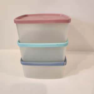 May include: Three rectangular food storage containers stacked on top of each other. The containers are translucent with colored lids and bands. The top lid is mauve, the middle band is turquoise, and the bottom band is blue.