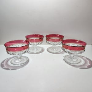 May include: Four clear glass champagne coupe glasses with a ruby red rim and a decorative pattern around the base. The glasses are arranged on a white surface, casting shadows.