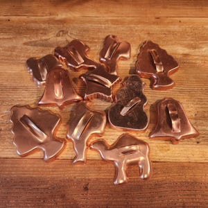 May include: A collection of copper-colored cookie cutters in various shapes, including a bell, tree, snowman, and camel. The cutters are arranged on a wooden surface, showcasing their detailed designs and metallic finish.