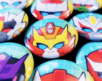 May include: A collection of colorful cartoon character buttons featuring Transformers characters. The buttons are round and have a shiny finish. The characters are depicted with large eyes and smiling expressions.