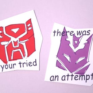 Op de afbeelding: Twee handgemaakte papieren uitsparingen met Transformers-logo's. Eén is rood met de tekst "your tried", en de andere is paars met de tekst "there was an an attempt".
