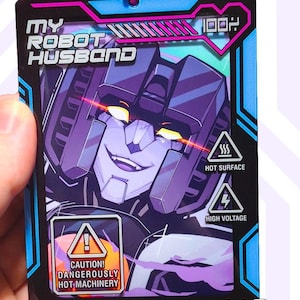 Könnte beinhalten: Ein rechteckiger Acryl-Anhänger mit einem lila Robotercharakter mit leuchtend gelben Augen und einem Grinsen. Das Design enthält den Text MY ROBOT HUSBAND, 100%, HOT SURFACE, HIGH VOLTAGE und CAUTION! DANGEROUSLY HOT MACHINERY. Mit rosa Kugelkette.