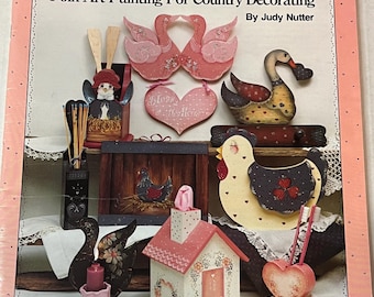 Puede incluir: Una portada de libro titulada "The Simple Joys" que presenta proyectos de pintura de arte popular. La portada muestra varias piezas de arte popular de madera, incluyendo cisnes, un corazón, una gallina y una casa, todo en un estilo de decoración campestre.