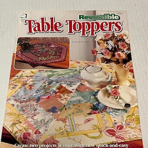 May include: A book titled "Reversible Table Toppers" by Anita Murphy. The book cover features a colorful patchwork table topper with a floral design. The book cover also includes a photo of a hexagonal table topper with a red and gold floral design.