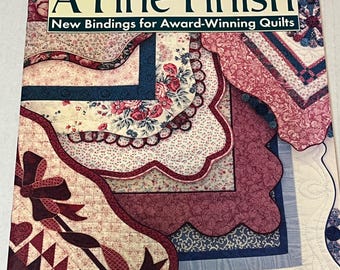 以下が含まれることがあります： 「A Fine Finish」というタイトルの本で、「New Bindings for Award-Winning Quilts」というサブタイトルが付いています。表紙は、赤、ピンク、青の色合いで、花柄と幾何学模様のパッチワークキルトのデザインが特徴です。著者の名前、Cody Mazuran が表示されています。