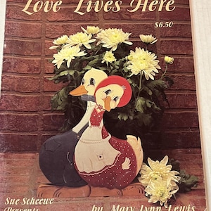 May include: A book titled "Love Lives Here" by Mary Lynn Lewis, featuring a painted wooden couple of ducks in overalls and a dress, surrounded by white mums. The book cover is brown with a brick pattern and the text "Sue Scheewe Presents" is at the bottom.