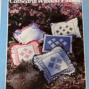 以下が含まれることがあります： キャシードラルウィンドウ枕のパターン。画像には、赤、白、青など、さまざまな色とデザインの完成した枕がいくつか表示されています。「A Patchwork Pattern and Instructions by Yours Truly」と書かれています。