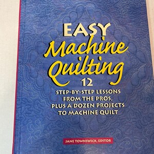 May include: A blue book with the title "Easy Machine Quilting 12" and the subtitle "Step-by-step lessons from the pros, plus a dozen projects to machine quilt". The book cover features a quilted design in blue and white.