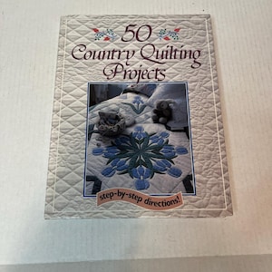 Pode incluir: Um livro intitulado "50 Country Quilting Projects" com uma capa acolchoada. A capa apresenta uma imagem de um edredom com desenhos florais azuis e brancos. O texto "step-by-step directions!" também é visível.
