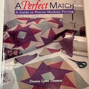 May include: A book titled "A Perfect Match: A Guide to Precise Machine Piecing" by Donna Lynn Thomas. The cover features a quilt with a star pattern in shades of purple, pink, and beige. The book is part of the "The Joy of Quilting" series.
