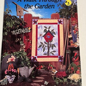 May include: A book titled "A Walk Through the Garden" with a number 3 on the cover. The cover features a colorful quilt with a birdhouse and a garden scene. The book is by Piecemakers.