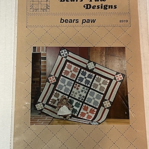 Könnte beinhalten: Eine Quilt-Schnittmusterpackung von Bears Paw Designs. Das Muster zeigt einen Quilt mit Bärenpfoten-Designs in Rot, Blau und Grau. Eine kleine Puppe ist ebenfalls zu sehen.