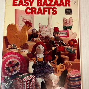 Puede incluir: Una portada de libro titulada "Easy Bazaar Crafts" con una ilustración colorida de artesanías hechas a mano, que incluye una muñeca de trapo pelirroja, una jirafa de madera, un gato de madera, un gallo de madera y una caja de rayas rojas y blancas.