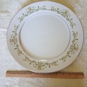 May include: A white ceramic dinner plate with a green and yellow floral design around the rim. The plate has a gold trim.