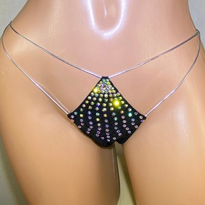 Puede incluir: Lingerie negra con un panel frontal en forma de triángulo cubierto de pedrería iridiscente. La lencería está sujeta por finas tiras plateadas.