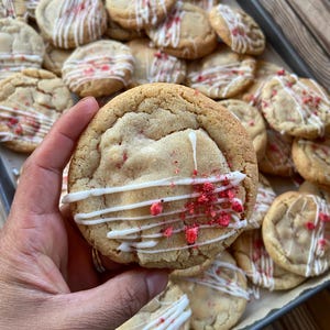 May include: A round, golden-brown baked cookie topped with a drizzle of white icing and small pieces of red dried fruit. The cookie has a soft, textured surface and is part of a larger batch of similar cookies arranged on a baking sheet.