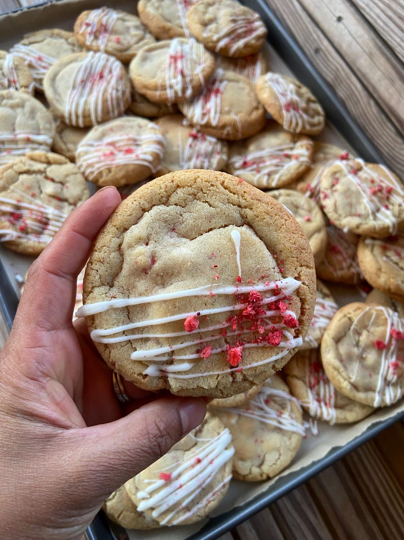May include: A round, golden-brown baked cookie topped with a drizzle of white icing and small pieces of red dried fruit. The cookie has a soft, textured surface and is part of a larger batch of similar cookies arranged on a baking sheet.