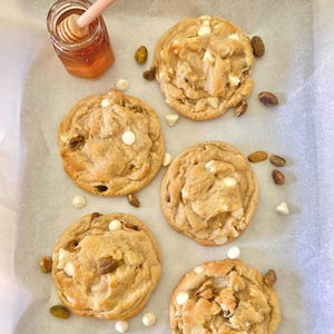 May include: Five round, golden-brown cookies with visible white chocolate chips and green pistachio pieces, arranged on parchment paper. A small glass jar filled with amber honey and a wooden honey dipper sits in the upper left corner.