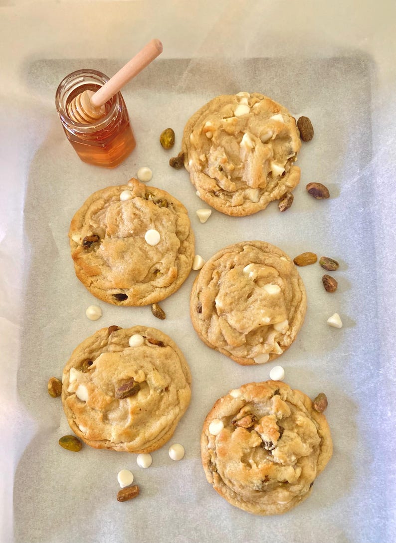 May include: Five round, golden-brown cookies with visible white chocolate chips and green pistachio pieces, arranged on parchment paper. A small glass jar filled with amber honey and a wooden honey dipper sits in the upper left corner.