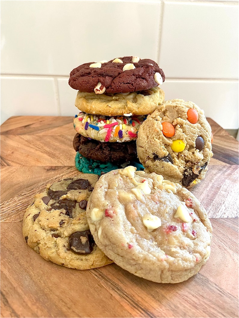 May include: A stack of assorted cookies including a dark chocolate cookie with white chips, a sugar cookie with sprinkles, a dark chocolate cookie, and a teal cookie. Beside the stack are a chocolate chip cookie and a white chip cookie.