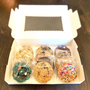 May include: A white cardboard box containing nine individually wrapped round cookies arranged in three rows. The cookies feature various textures and colors, including a bright teal cookie with dark chocolate chips and a cookie covered in colorful sprinkles.