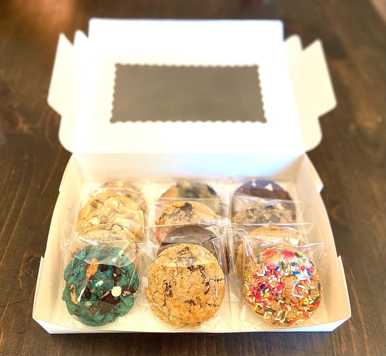 May include: A white cardboard box containing nine individually wrapped round cookies arranged in three rows. The cookies feature various textures and colors, including a bright teal cookie with dark chocolate chips and a cookie covered in colorful sprinkles.