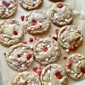 May include: A collection of round, baked cookies arranged on parchment paper. Each cookie features a light brown, textured surface embedded with bright red dried strawberry pieces and small, solid white chocolate chips throughout.
