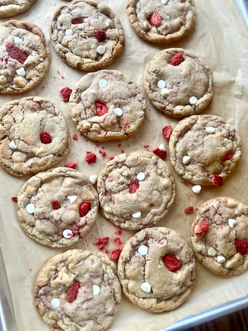 May include: A collection of round, baked cookies arranged on parchment paper. Each cookie features a light brown, textured surface embedded with bright red dried strawberry pieces and small, solid white chocolate chips throughout.