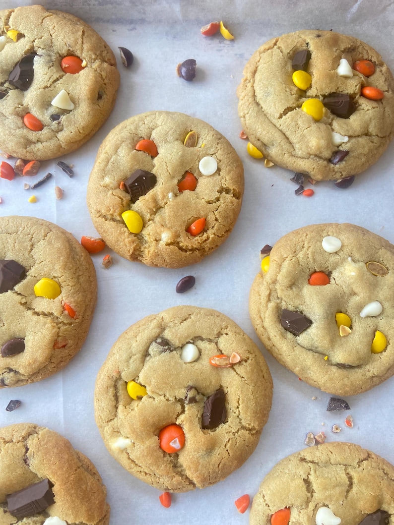 May include: Several round, golden-brown baked cookies are arranged on white parchment paper. Each cookie is studded with chunks of dark chocolate, white chocolate chips, and small, round, colorful candy-coated chocolate pieces in orange and yellow.