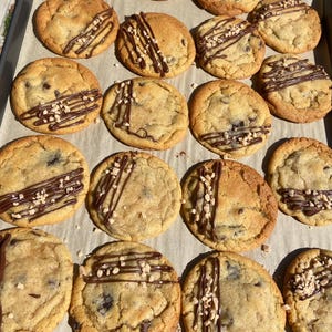 May include: A batch of sixteen round, golden-brown chocolate chip cookies arranged on parchment paper. Each cookie is drizzled with dark chocolate and topped with small, light-colored nut pieces, creating a textured, sweet baked treat.
