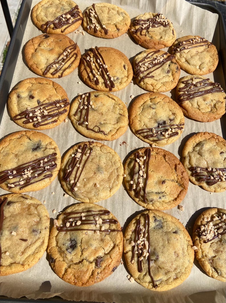 May include: A batch of sixteen round, golden-brown chocolate chip cookies arranged on parchment paper. Each cookie is drizzled with dark chocolate and topped with small, light-colored nut pieces, creating a textured, sweet baked treat.