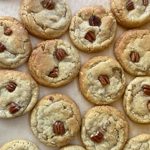 May include: A collection of round, golden-brown baked cookies arranged on parchment paper. Each cookie has a soft, textured surface with visible cracks and is topped with one or more whole pecan halves embedded into the dough.
