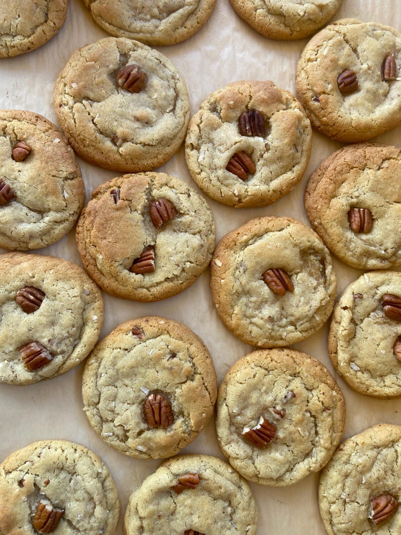May include: A collection of round, golden-brown baked cookies arranged on parchment paper. Each cookie has a soft, textured surface with visible cracks and is topped with one or more whole pecan halves embedded into the dough.