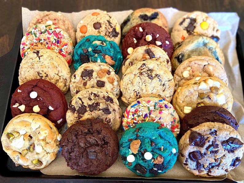 May include: A variety of round baked cookies arranged on a tray, featuring diverse textures and ingredients including chocolate chips, colorful sprinkles, white chocolate chunks, red velvet dough, and bright blue dough with various inclusions.