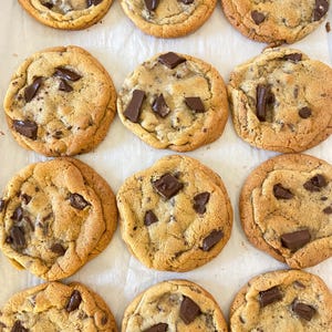 May include: Twelve round chocolate chip cookies arranged in a grid on white parchment paper. Each golden-brown cookie features a soft, baked texture with numerous dark brown chocolate chunks embedded throughout the surface of the dough.