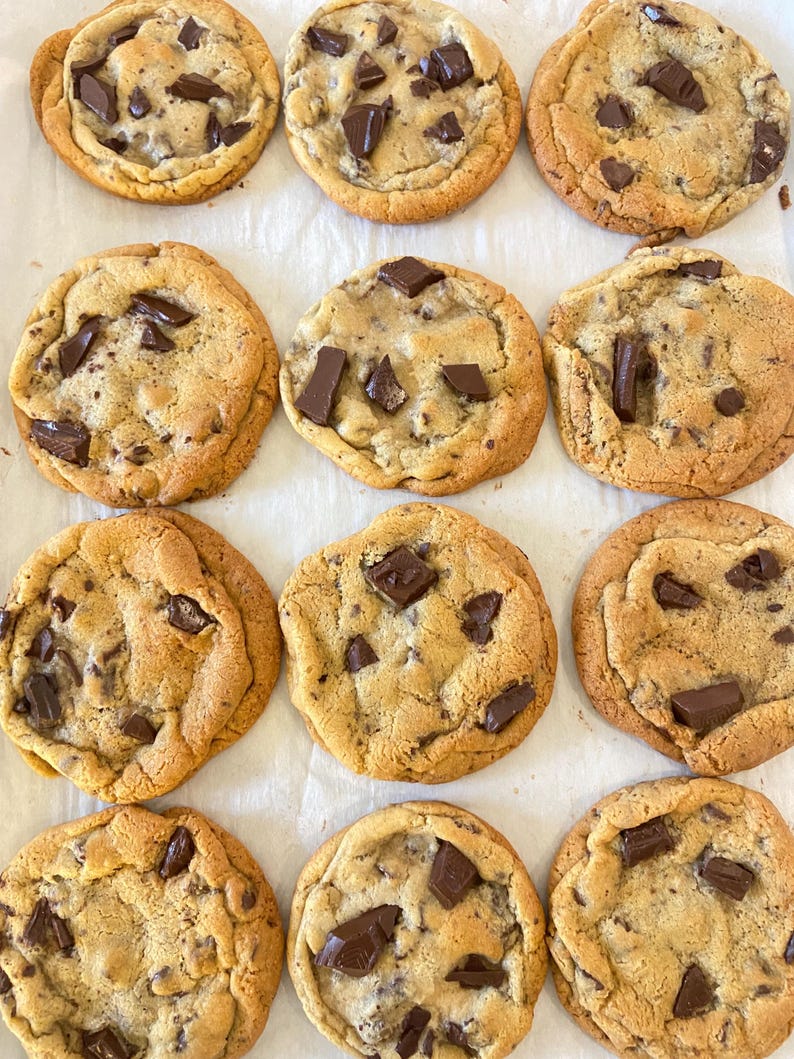 May include: Twelve round chocolate chip cookies arranged in a grid on white parchment paper. Each golden-brown cookie features a soft, baked texture with numerous dark brown chocolate chunks embedded throughout the surface of the dough.