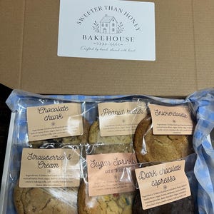 May include: A cardboard box containing six individually wrapped cookies with labels reading Chocolate Chunk, Peanut Butter, Snickerdoodle, Strawberries & Cream, Sugar Sprinkle, and Dark Chocolate Espresso, with a Sweeter Than Honey Bakehouse logo.