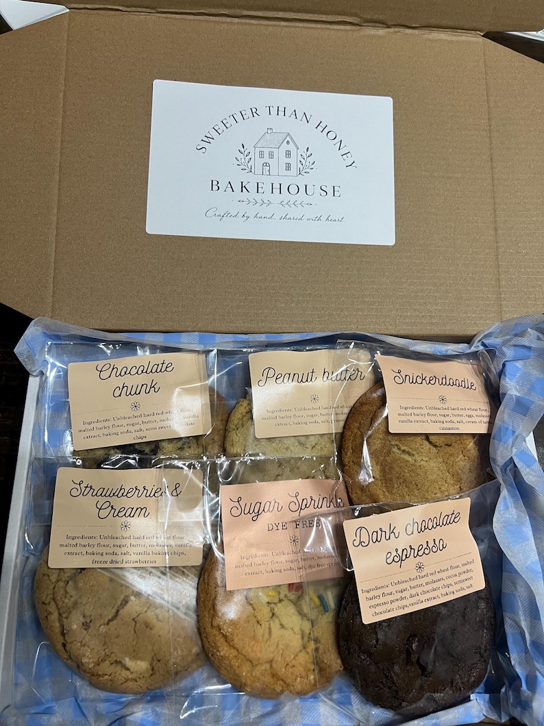 May include: A cardboard box containing six individually wrapped cookies with labels reading Chocolate Chunk, Peanut Butter, Snickerdoodle, Strawberries & Cream, Sugar Sprinkle, and Dark Chocolate Espresso, with a Sweeter Than Honey Bakehouse logo.