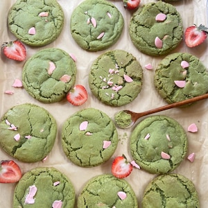 May include: A collection of round, green matcha cookies featuring pink chocolate chips and fresh strawberry slices. A small wooden spoon containing a mound of green matcha powder is positioned among the cookies on a sheet of parchment paper.